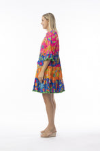 Load image into Gallery viewer, Orientique Mira Flores printed open neck tiered organic cotton dress displayed on a model, showcasing floral pattern and flowing design.