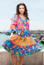Load image into Gallery viewer, Orientique Mira Flores printed open neck tiered organic cotton dress, featuring colorful floral patterns and flowing tiered design.
